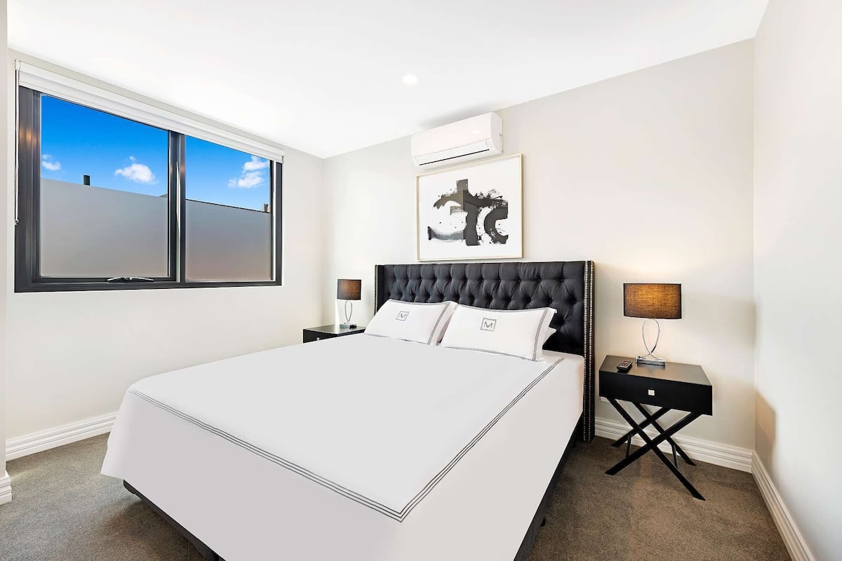 A hotel room with a large bed, two bedside tables with lamps, a window with blinds, an air conditioning unit, and a framed artwork on the wall.
