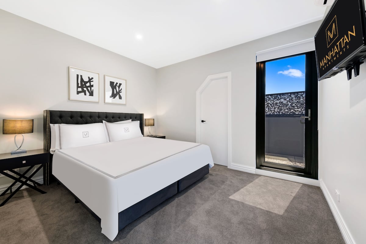 A modern bedroom with a large bed, a nightstand, a desk, and a sliding door leading to a balcony.