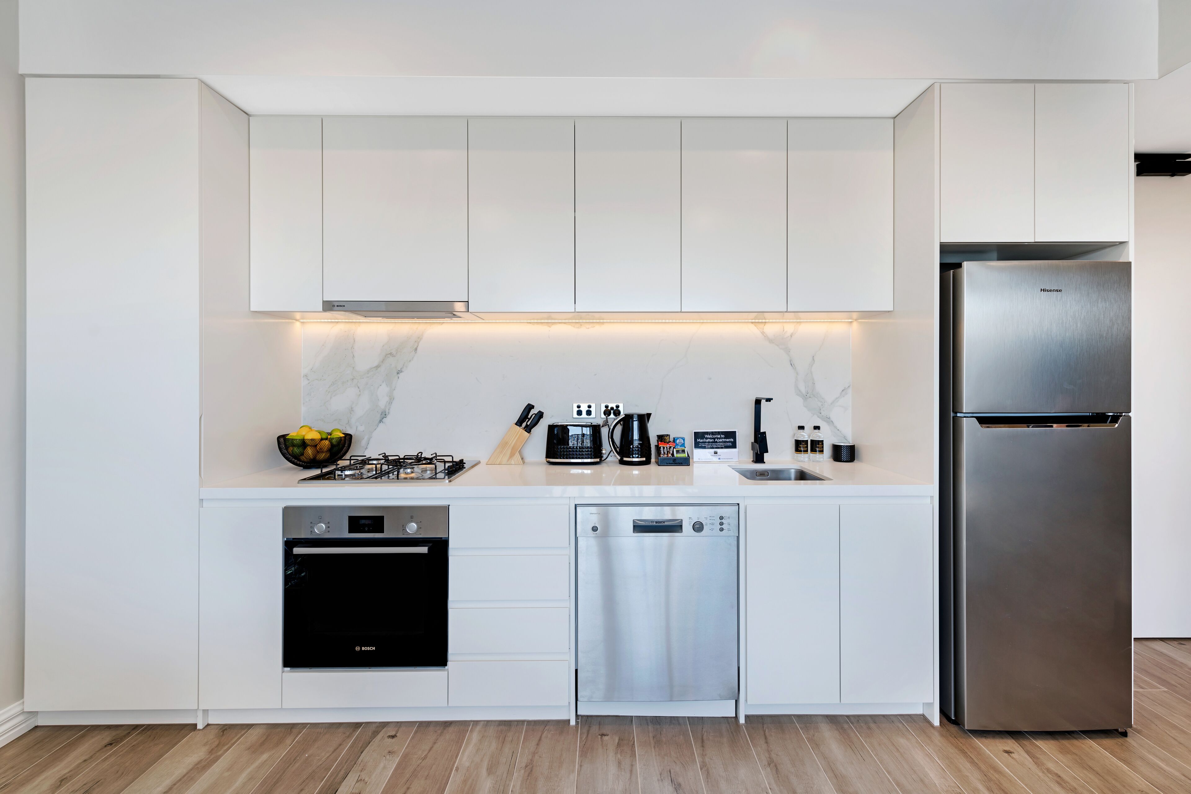 A modern kitchen with white cabinetry, a Bosch dishwasher, a Bosch oven, a Bosch coffee maker, and a stainless steel refrigerator.