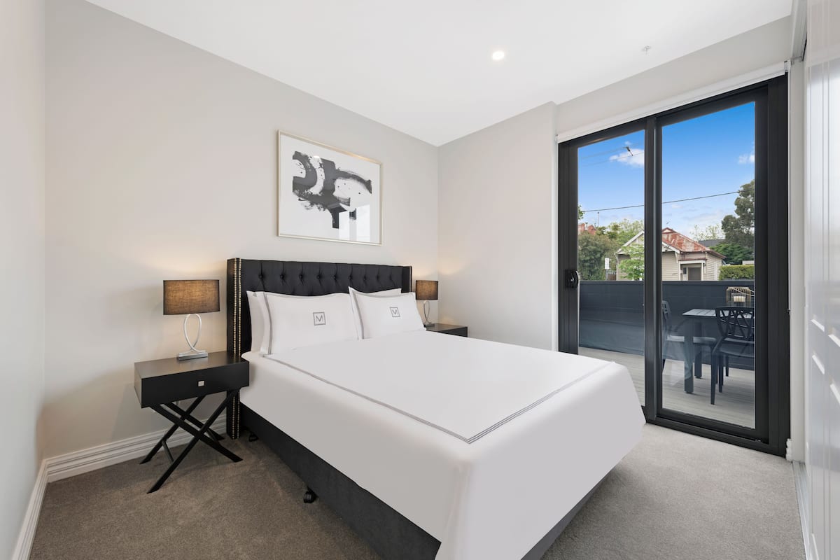 A modern bedroom with a large bed, a nightstand, a wall-mounted abstract painting, and a sliding glass door leading to an outdoor area.