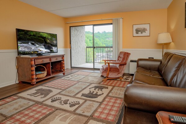 Un salon comprenant un téléviseur, un canapé, un fauteuil, un tapis orné d’empreintes de pattes et une vue sur l’extérieur, visible à travers une porte vitrée.