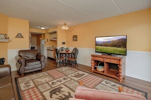 A living room with a television, a sofa, a dining table with chairs, and a kitchen in the background.