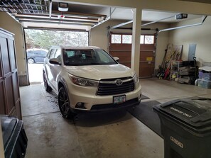 Un VUS blanc est garé dans un garage dont le sol est en béton, qui est équipé d’une porte de garage et où se trouve une poubelle portant l’inscription « Déchets uniquement ».
