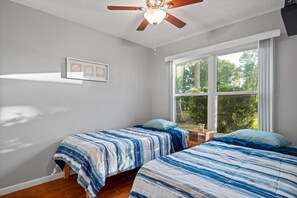 Un dormitorio con dos camas, un ventilador de techo y un ventanal con vistas a la vegetación.