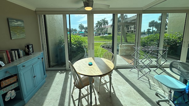 Una habitación iluminada por el sol con una mesa y sillas, un ventilador de techo, un ventanal corredero a un jardín y un armario azul con un letrero que dice 'Por favor, quitar'.