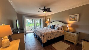 Un dormitorio con cama, ventilador de techo, televisión, escritorio con lámpara y vistas al exterior.
