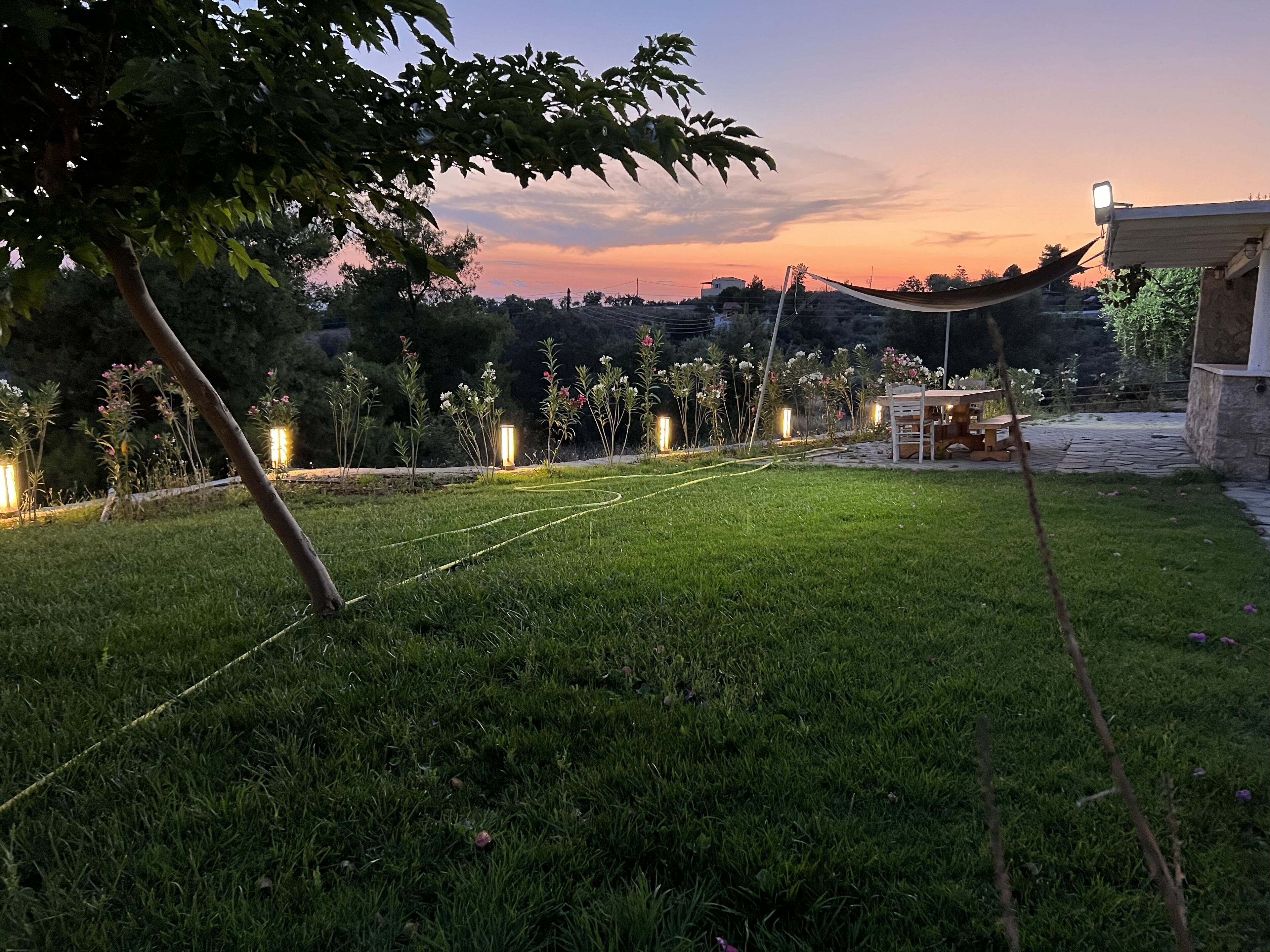 Uma área externa gramada com mesa e cadeiras de madeira, cercada por plantas e iluminada por luzes de cordão.