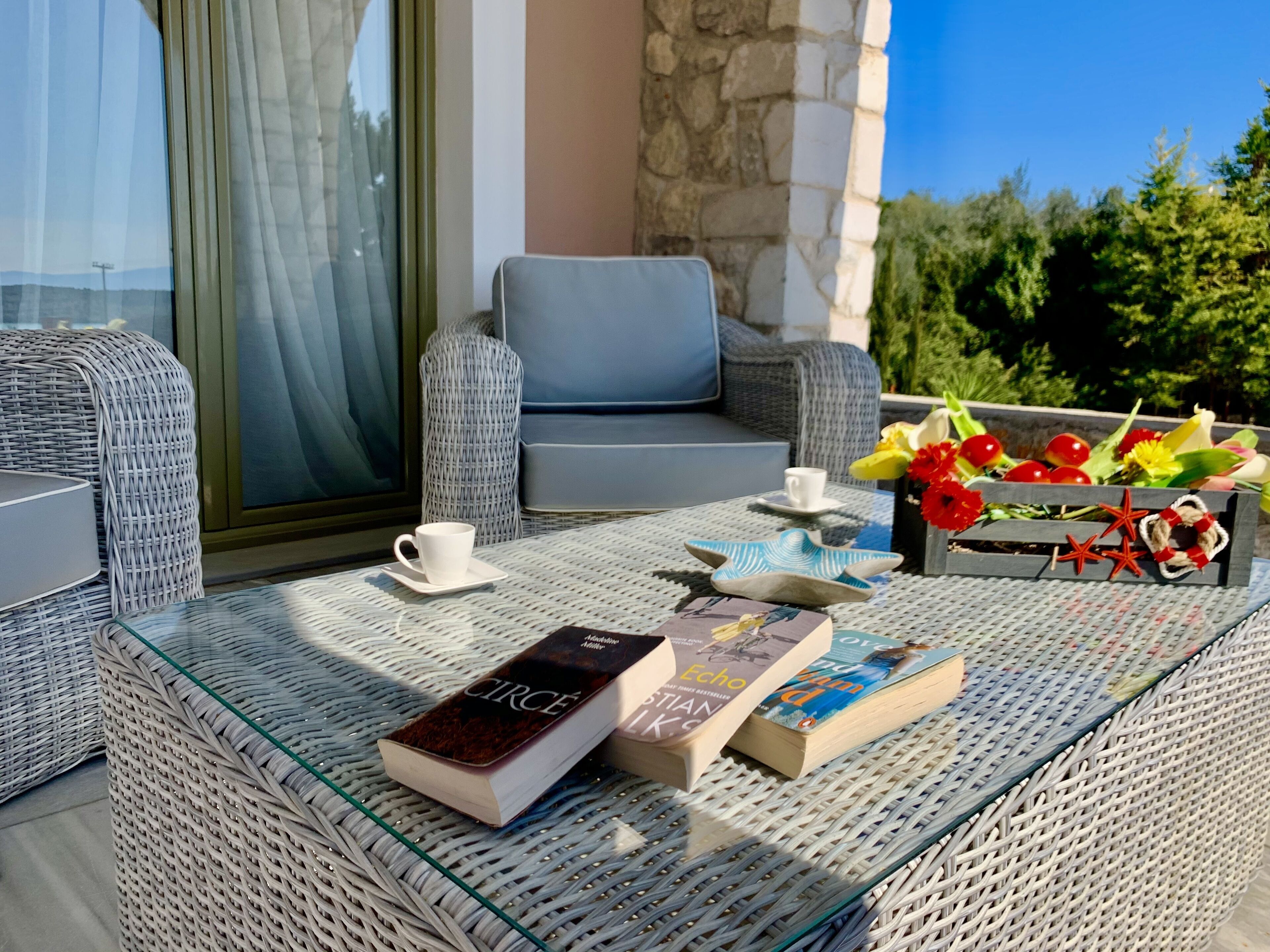 Un balcón con mobiliario de mimbre, una mesa de vidrio, libros, una taza y flores.