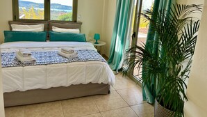 A bedroom with a bed, pillows, a nightstand, a lamp, a window with curtains, and a potted plant. - Villa Victory Porto Heli (Ermionida)