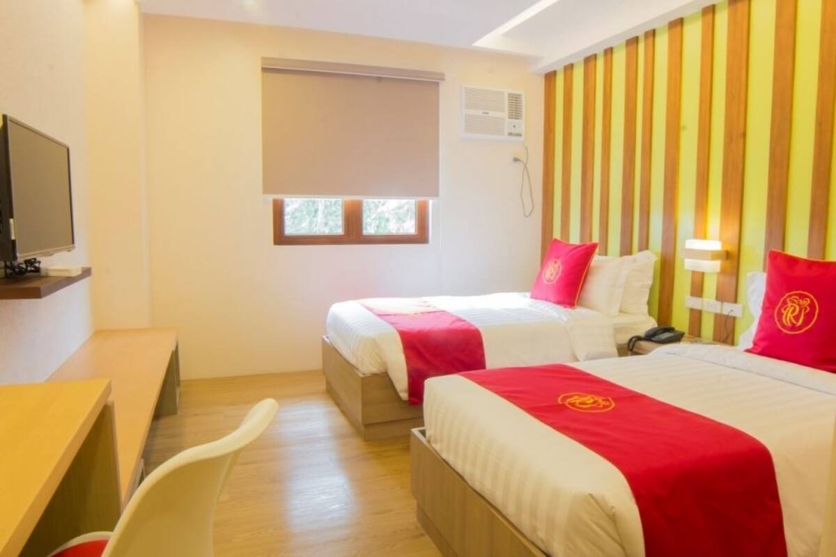 A hotel room with two beds, a desk, a TV, and a window with blinds.