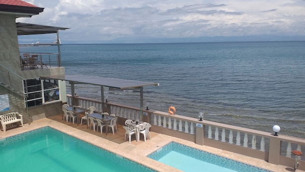 A pool area with a view of the sea, outdoor seating, and a building with a red roof. - Ocean Breeze Resort (Toledo City)