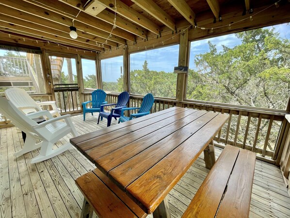 A wooden deck with a table and chairs, large windows, and a view of trees.