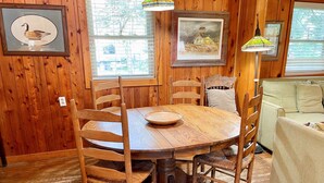 A wooden dining table with chairs, framed pictures on the walls, and a window with blinds. - Happy Place:  Pet friendly, fenced rear yard with deck. (Ocracoke)
