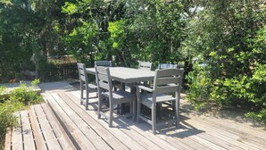 A wooden deck with a gray table and chairs, surrounded by lush greenery. - Happy Place:  Pet friendly, fenced rear yard with deck. (Ocracoke)