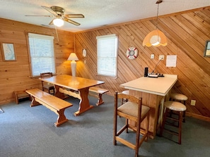 A room with a wooden interior, a ceiling fan, a table with benches, and a small bar area with stools.
