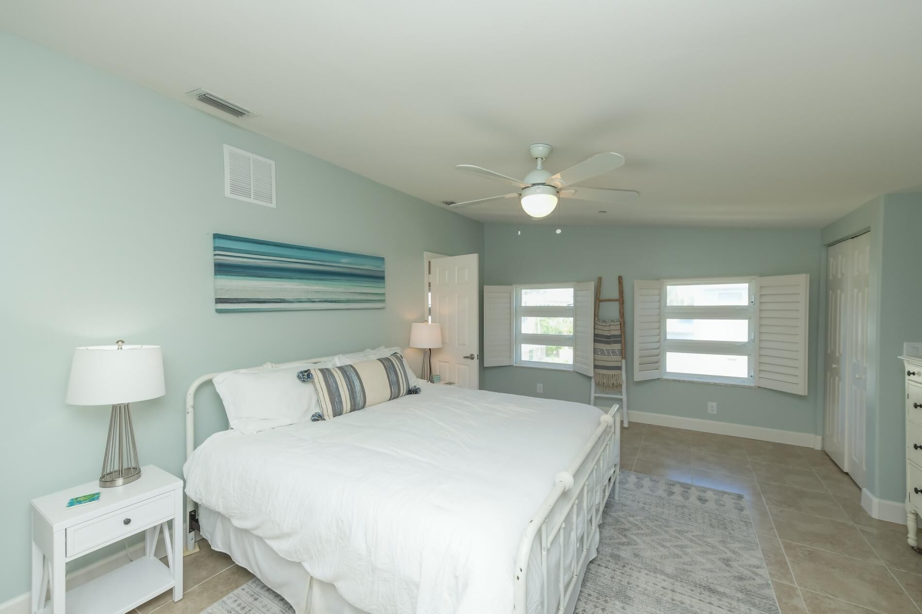A bedroom with a bed, a nightstand, a lamp, a ceiling fan, and a window with shutters.