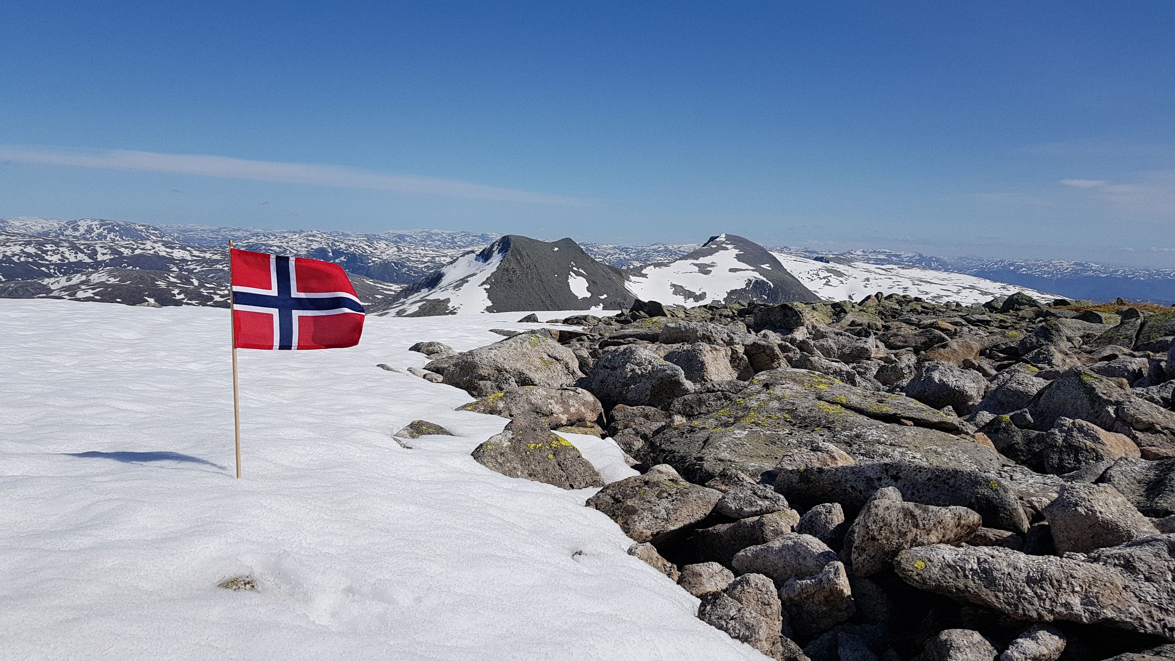 A snow-covered mountain peak with a flag, rocky terrain, and distant snow-capped mountains.