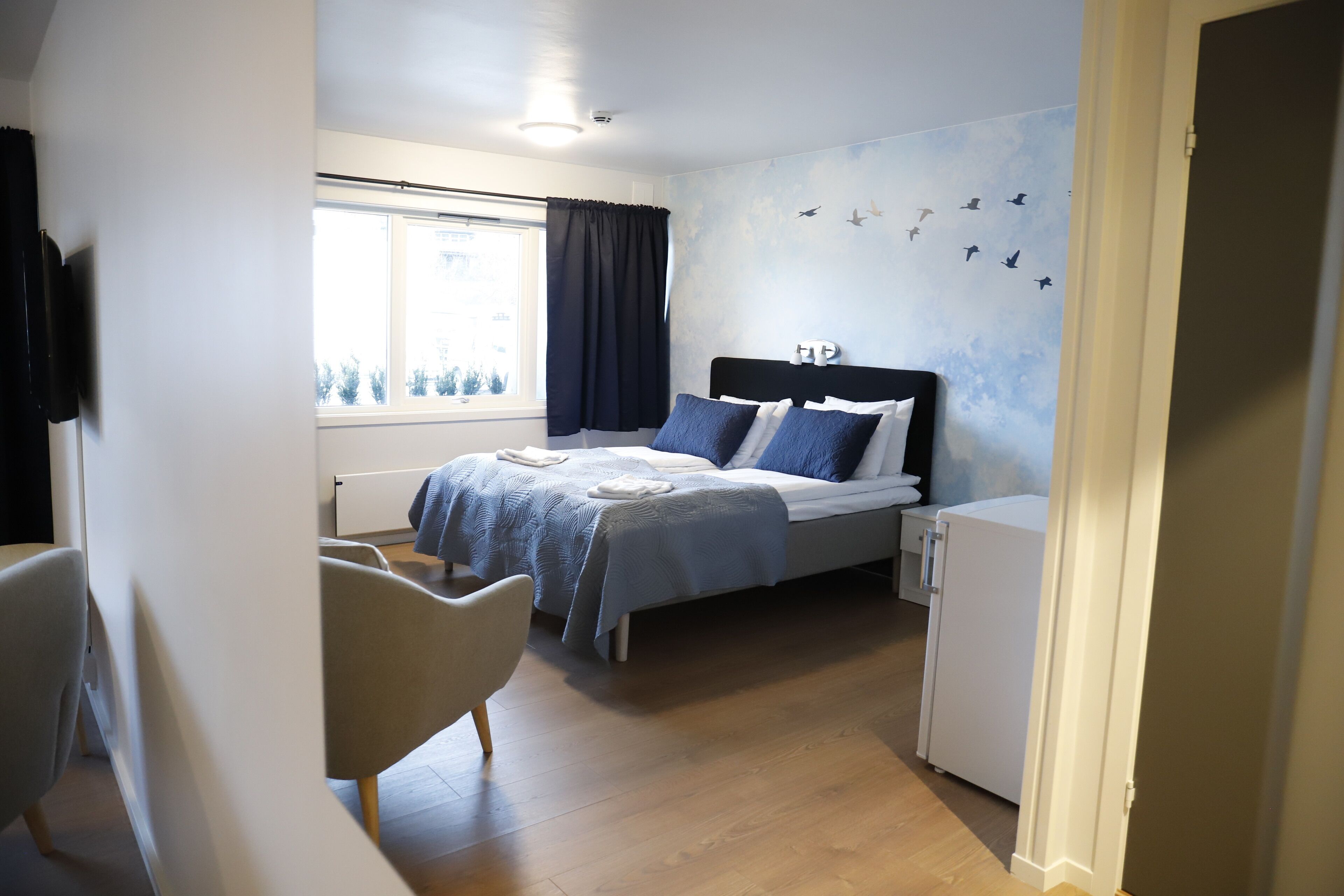 Quarto com cama, uma cadeira, uma mesinha, janela com cortinas e parede com adesivos de pássaros.