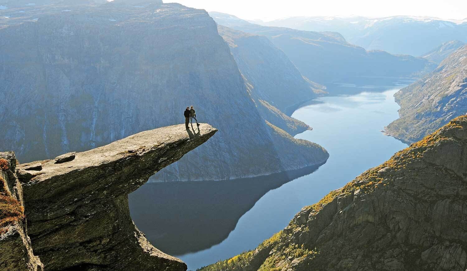 A person standing on a narrow ledge overlooking a deep valley with a lake.