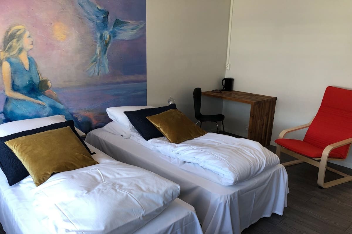 A room with two beds, a red chair, a small table, and a mural on the wall.