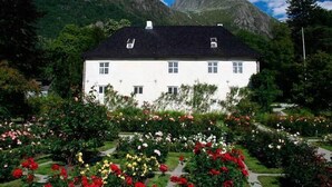 A white building with a black roof, surrounded by a garden with various flowers and a mountainous backdrop. - Husnes Sentrum Hotell (Kvinnherad)