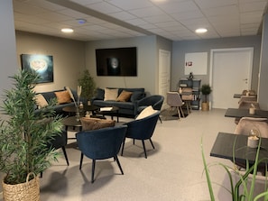 A modern living room with a sofa set, coffee table, and chairs, a flat-screen TV mounted on the wall, and a small bookshelf.