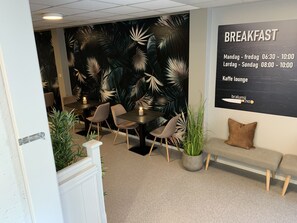 A breakfast area with tables and chairs, a wall with a tropical leaf mural, and a sign indicating breakfast hours. - Husnes Sentrum Hotell (Kvinnherad)