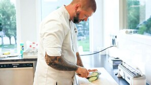 A person in a white chef's coat cutting a cucumber on a kitchen counter with a window in the background. - Husnes Sentrum Hotell (Kvinnherad)