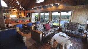 A cozy living area with a sofa, chairs, and a coffee table. There is a kitchen area with cabinets and a dining table. Large windows offer a view of the outdoors. - Cuttin' Sage:  Tropical flavor, reverse floorplan, canal, marsh views. (Ocracoke)