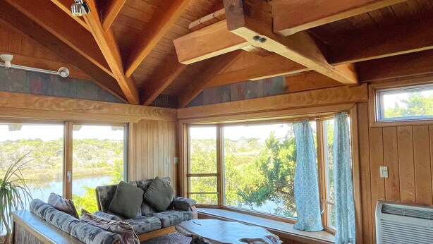 A cozy room with a wooden ceiling, large windows, a sofa, a table, and a view of greenery outside. - Cuttin' Sage:  Tropical flavor, reverse floorplan, canal, marsh views. (Ocracoke)