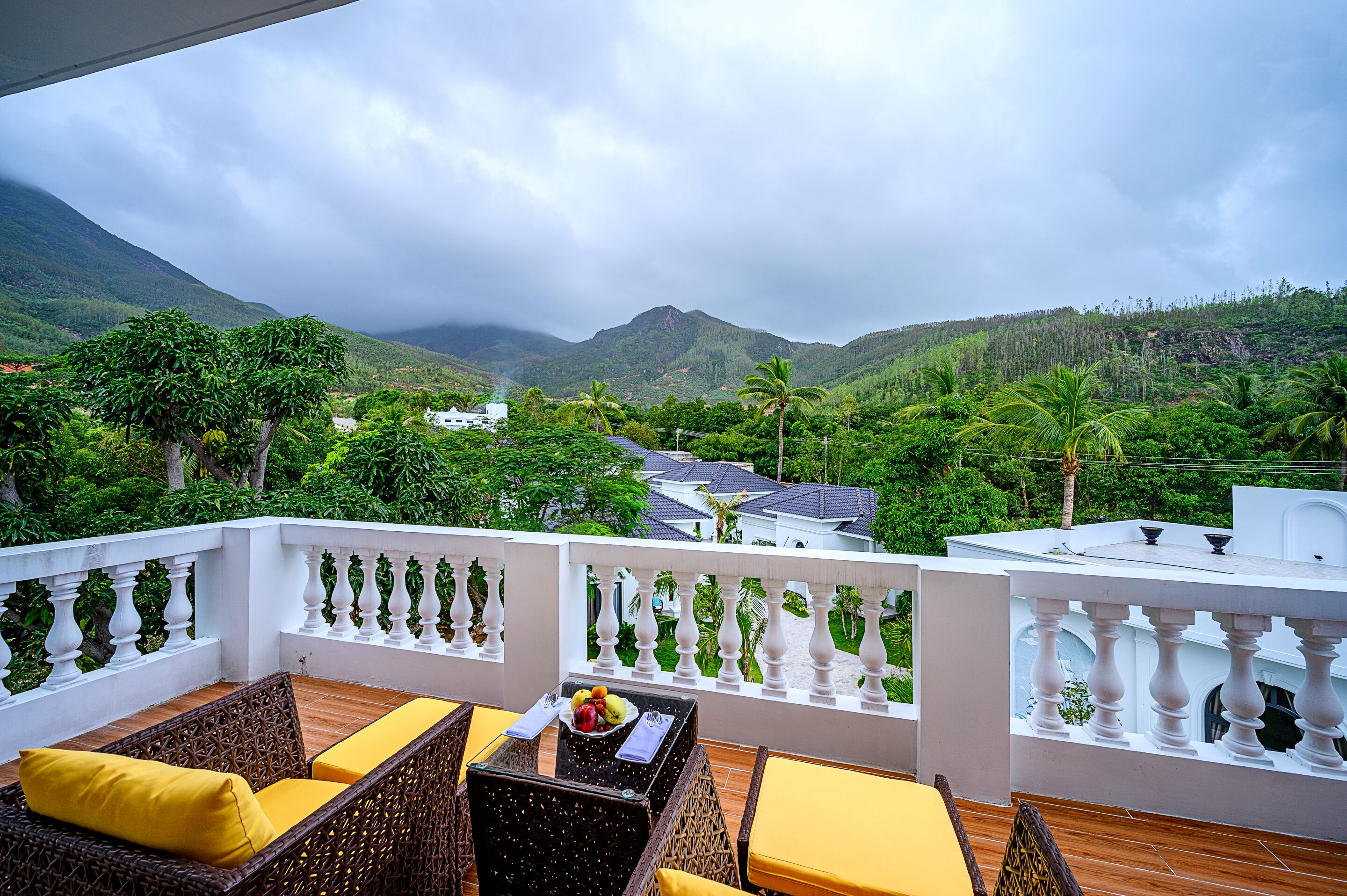 A balcony with wicker furniture and yellow cushions, overlooking a lush valley with mountains and a white building in the distance.