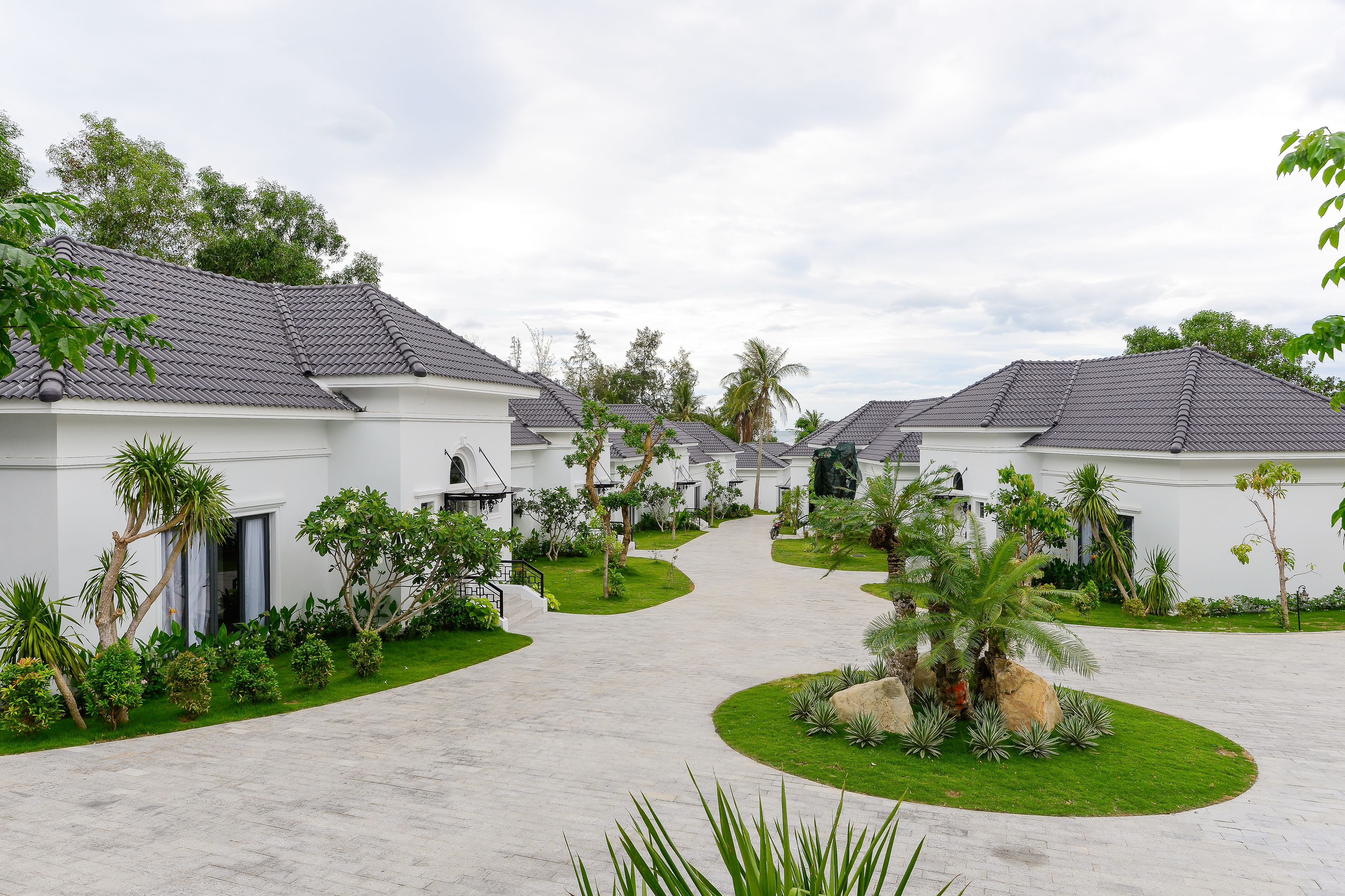 Un barrio residencial con casas blancas, senderos pavimentados y abundante vegetación, incluyendo palmeras y arbustos.