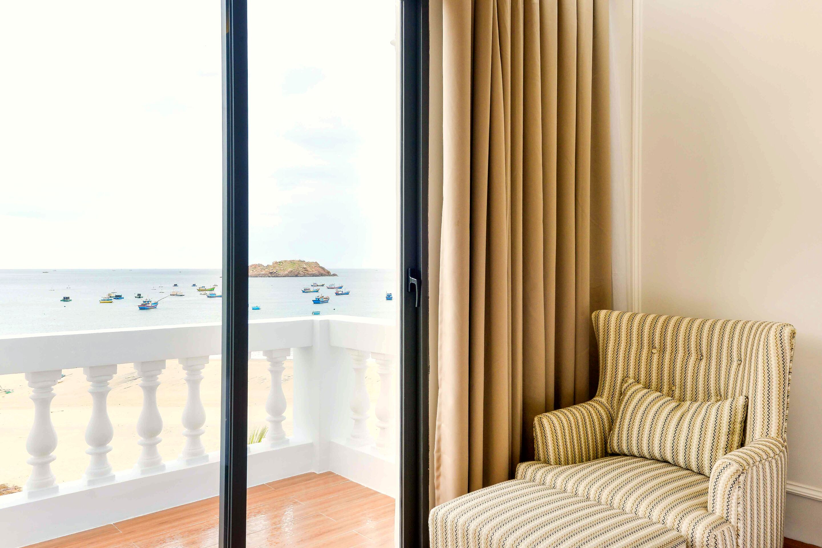 A balcony with a view of the sea and boats, a striped armchair, and a white balustrade.