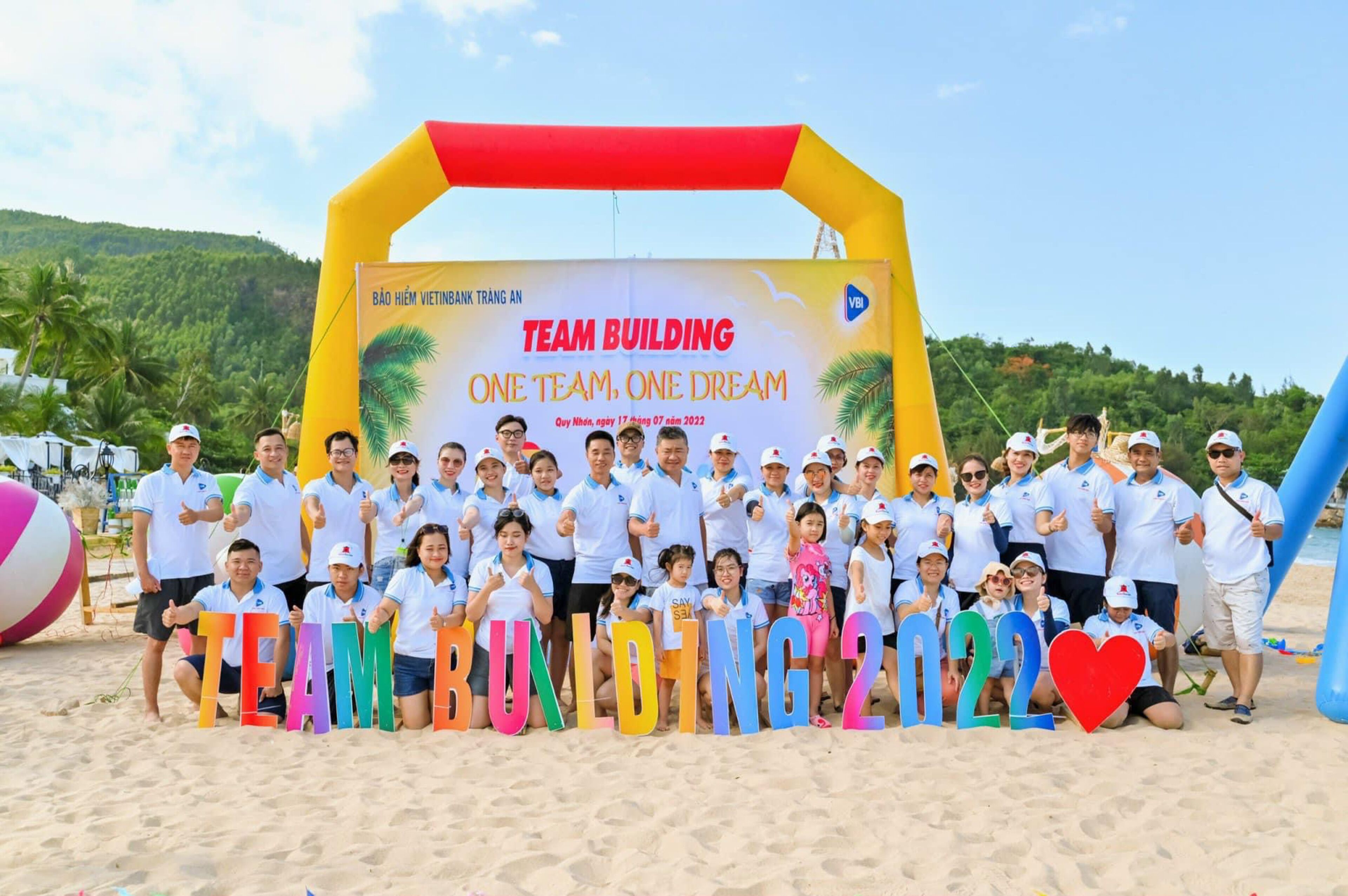 Eine Gruppe von Menschen posiert vor einem bunten, aufblasbaren Bogen mit einem Banner, auf dem steht: TEAM BUILDING ONE TEAM ONE DREAM.