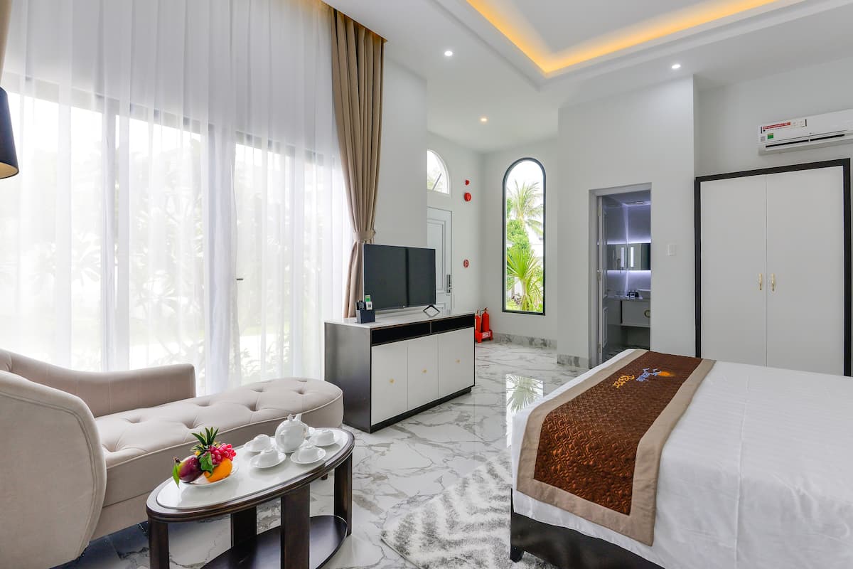 Ein Hotelzimmer mit Bett, Sofa, Fernseher und Esstisch mit Obst, durch einen Bogen hindurch ist das Badezimmer zu sehen.