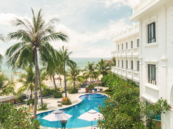 A pool area with palm trees, a view of the ocean, and a white building with balconies. - Seaside Boutique Resort Quy Nhon (Song Cau)