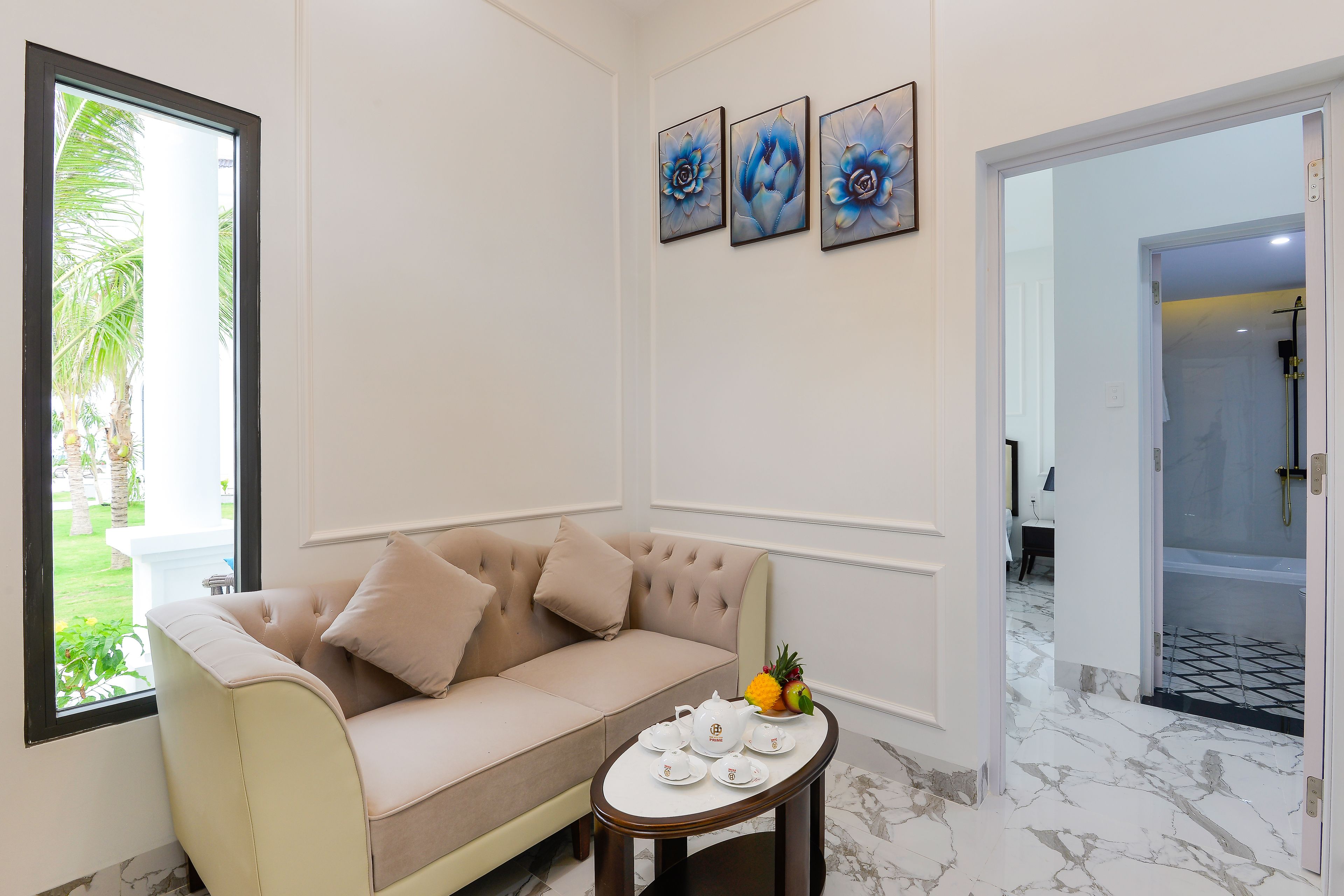A room with a beige sofa, a small table with a fruit arrangement, and three framed artworks on the wall.