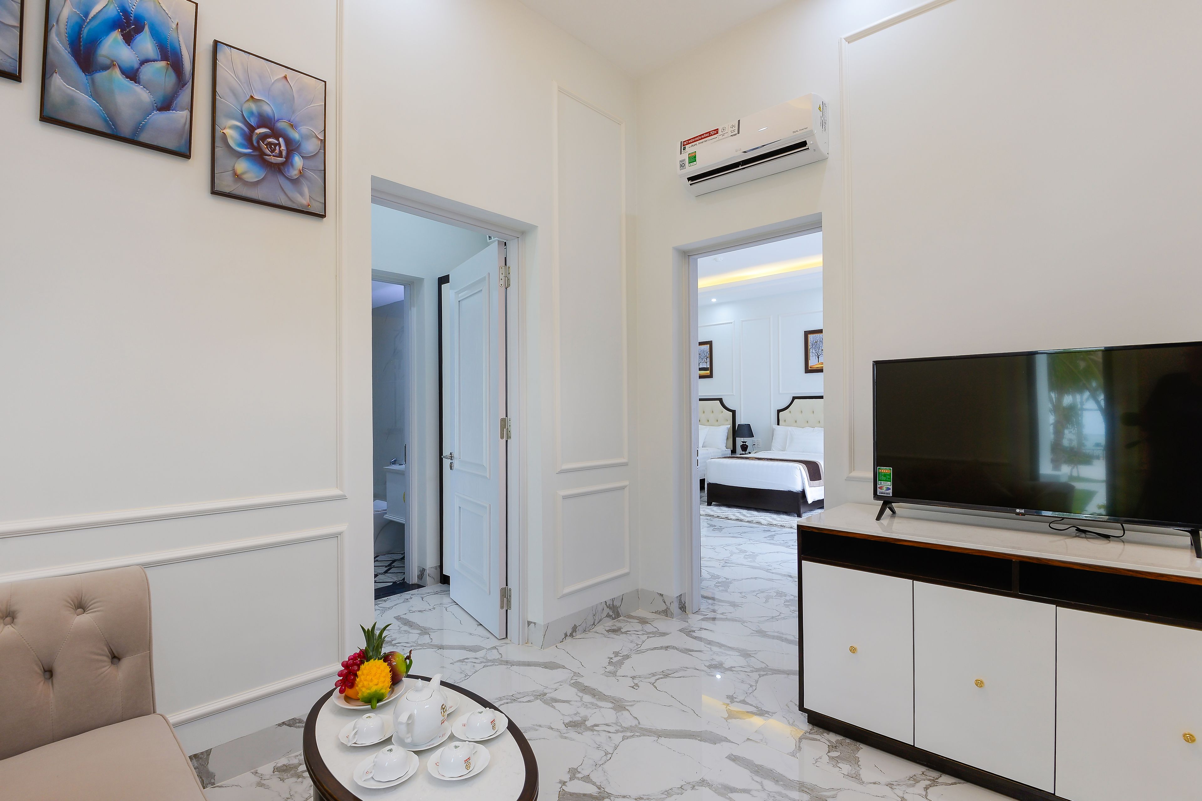 A hotel room with a flat-screen TV, a marble floor, a dining table with plates and a pineapple, and artwork on the wall.