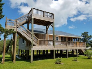 A two-story wooden deck with a staircase leading to a balcony, surrounded by a grassy area and trees.