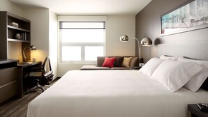 A hotel room with a large bed, a desk with a chair, a bookshelf, and a window providing natural light. - Hyatt House Indianapolis / Fishers (Fishers)