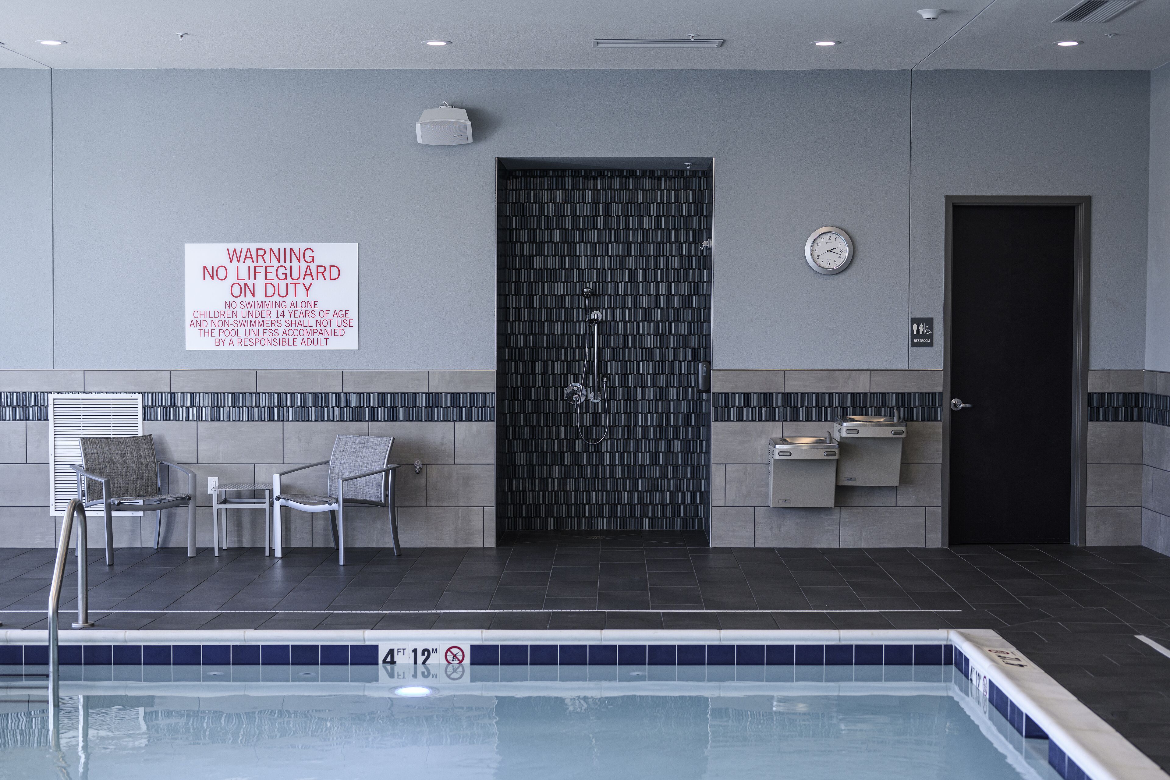 Une zone autour d’une piscine, avec un panneau d’avertissement indiquant la présence d’un sauveteur, une porte et une horloge au mur.
