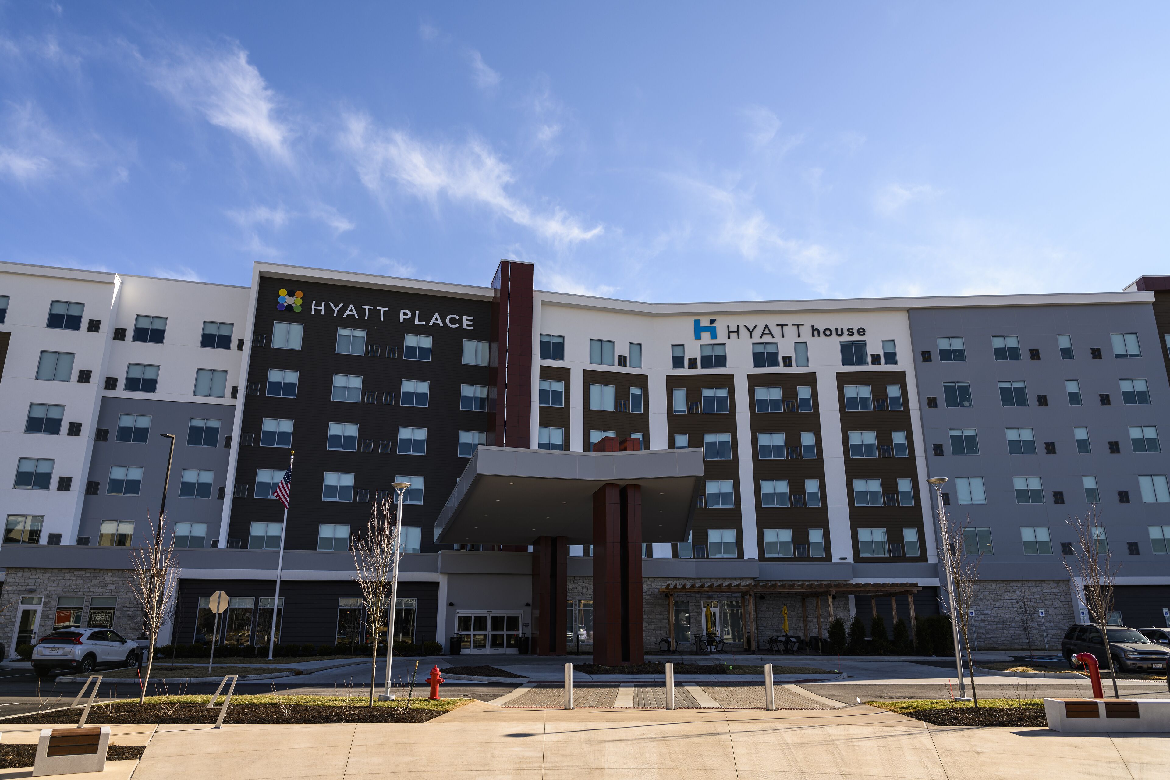 A modern hotel building with multiple floors, a prominent entrance, and a landscaped area with trees and a fire hydrant.