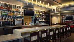 A bar area with multiple shelves of bottles, a long counter, and several bar stools. - Hyatt Place Indianapolis / Fishers (Fishers)