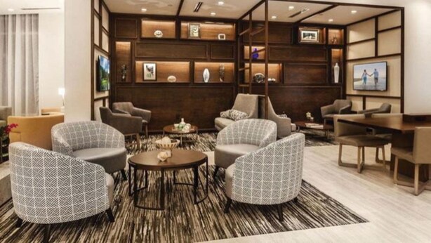 A modern lounge area with a central coffee table surrounded by patterned armchairs, a wooden accent wall with shelves, and a television screen. - Hyatt Place Indianapolis / Fishers (Fishers)