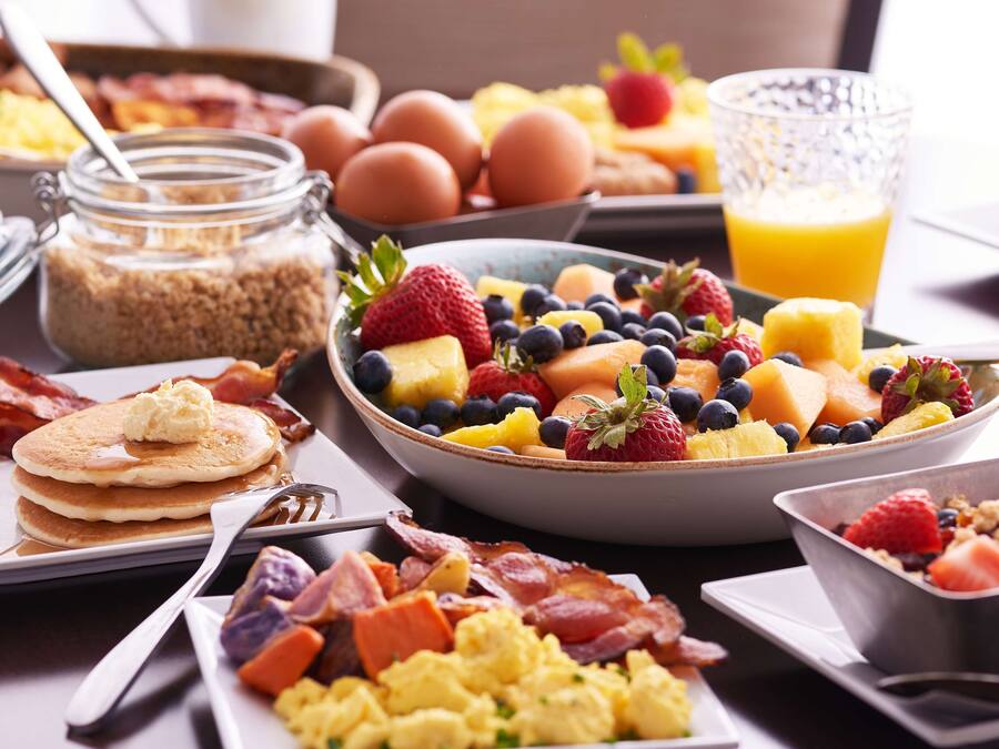 Ein Frühstücksbuffet mit verschiedenen Speisen, darunter Pfannkuchen, Rührei, Speck und einer Obstschale.