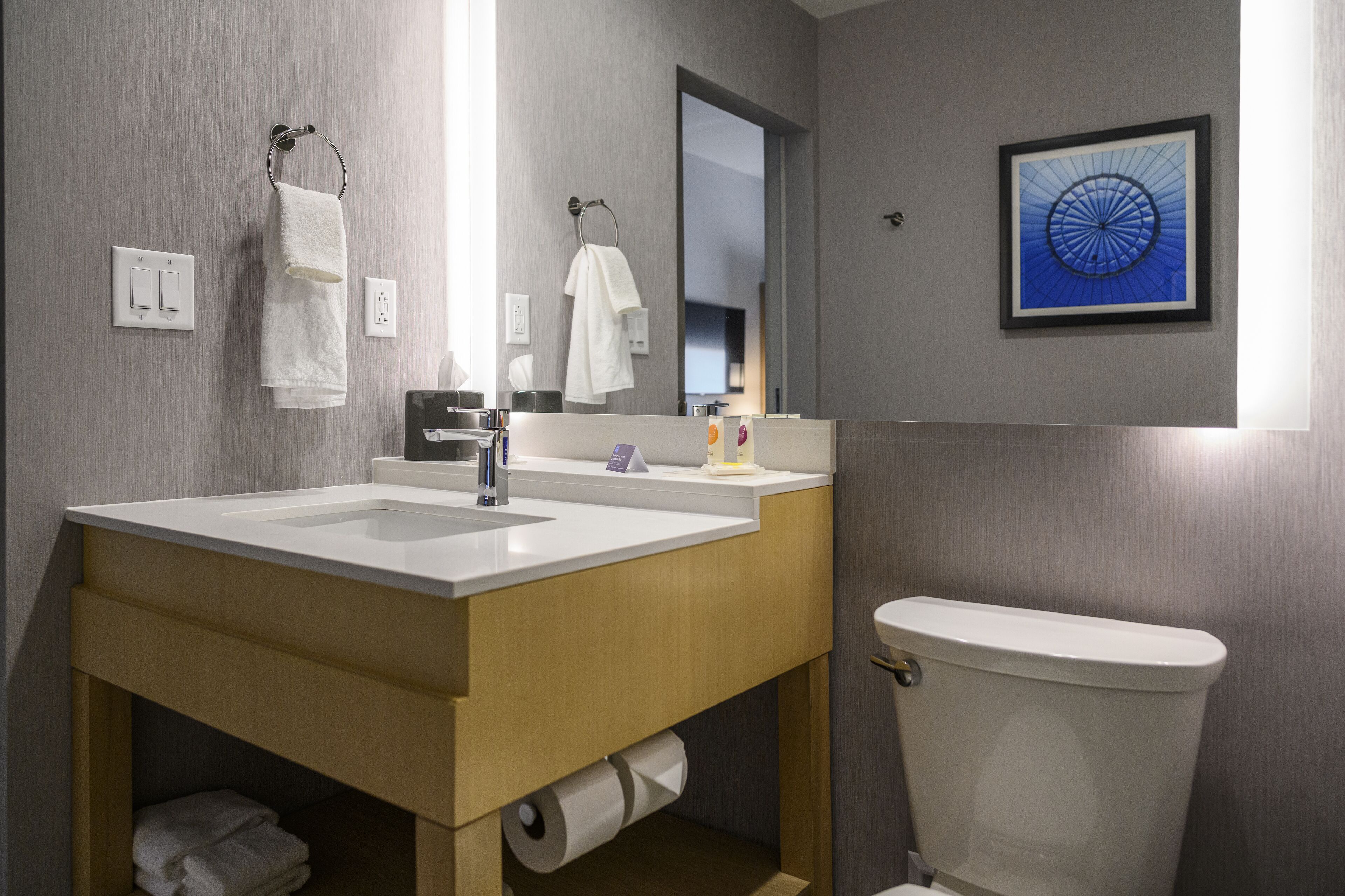 Et moderne badeværelse med hvid vask, spejl og toilet. To håndklæder hænger på kroge, et billede i ramme på væggen og toiletpapir under vasken.