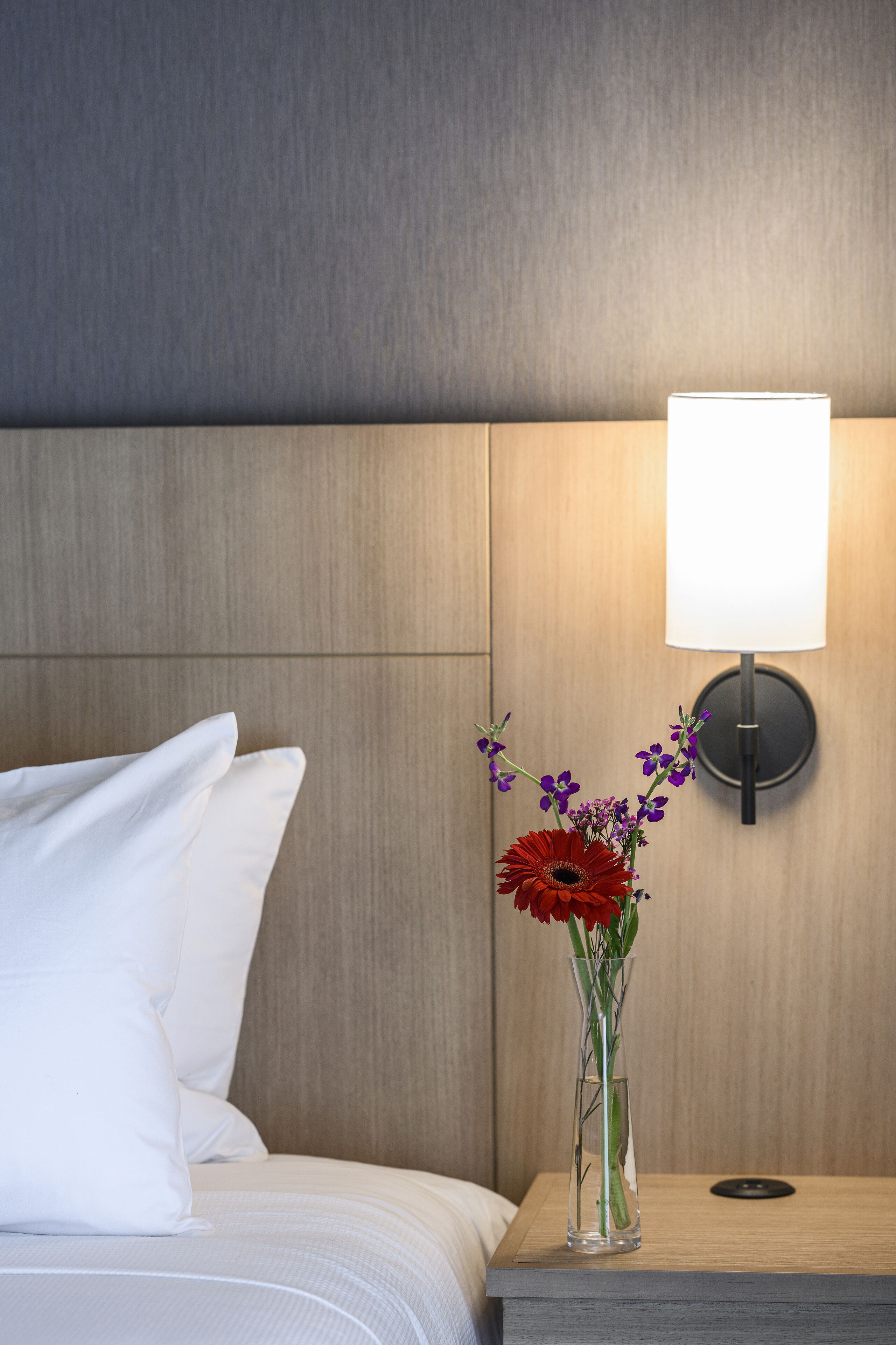 En pænt redt seng med hvide sengetøj og puder, en klar vase med blomster, og en vægmonteret lampe på et trænatbord.
