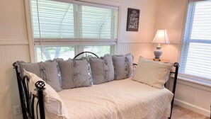 Een knus ingerichte kamer met een bed, een raam met zonwering, een wandlamp en een decoratief bord.