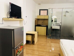 Quarto de hotel compacto com uma cama, uma pequena mesa, um frigorífico, uma televisão, uma kitchenette e uma casa de banho com cabine de duche de vidro.
