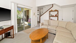 A living room with a sectional sofa, a wooden coffee table, a TV on a stand, a bookshelf, and a staircase. - South Shore Island Getaway at Poipu Crater 7 (Po'ipu)